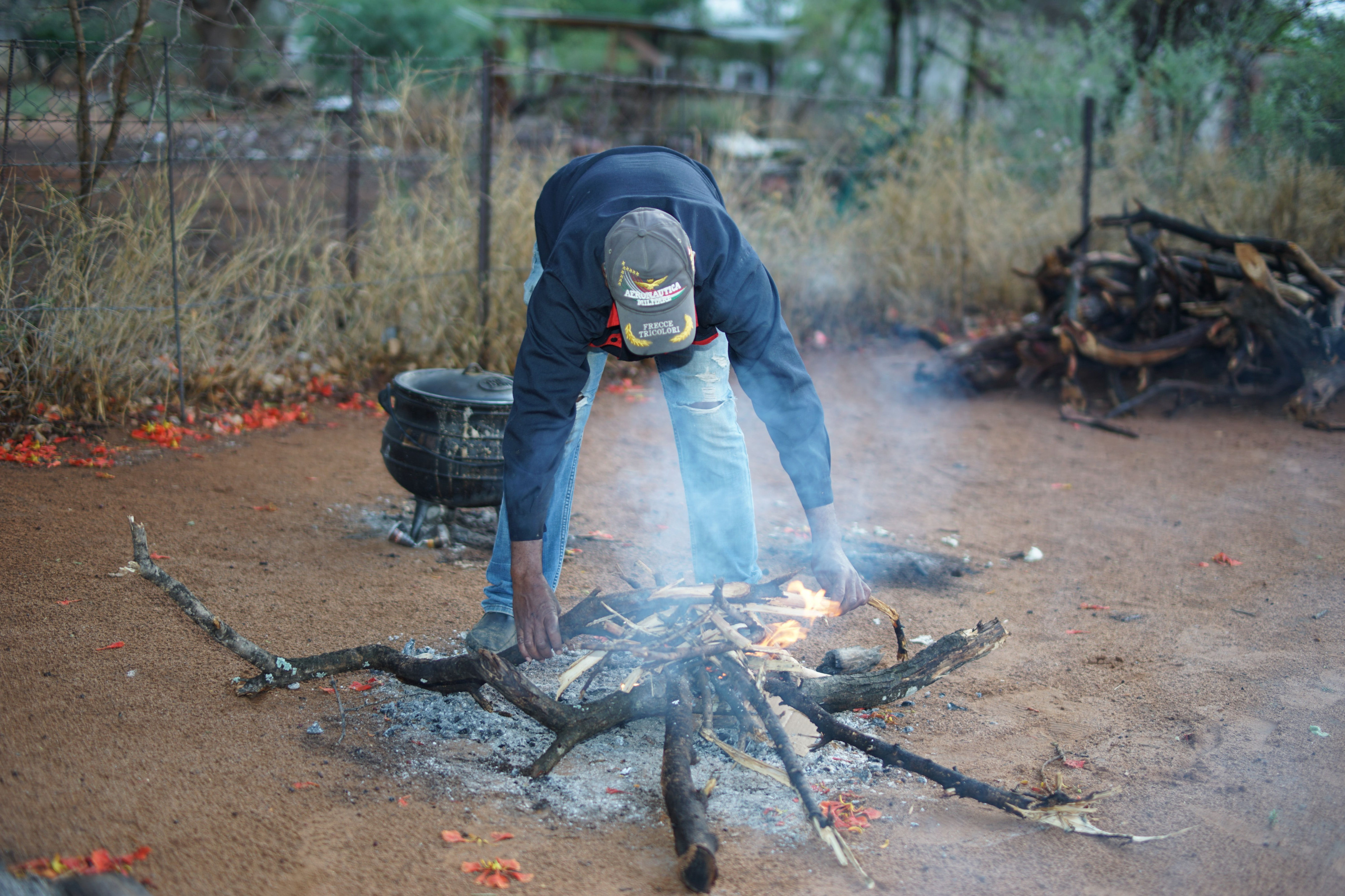 A man tends a small outdoor fire using traditional methods at the edge of a forest clearing.