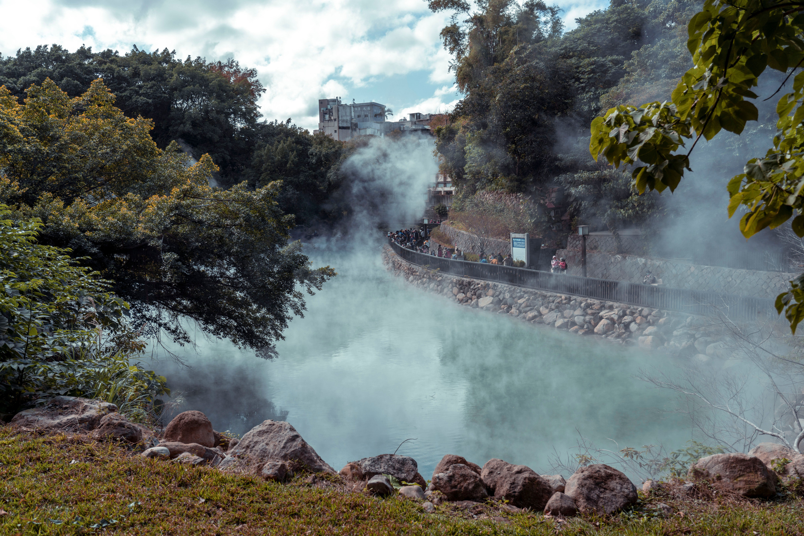 Steam rises from a hot spring surrounded by dense tropical forest greenery.