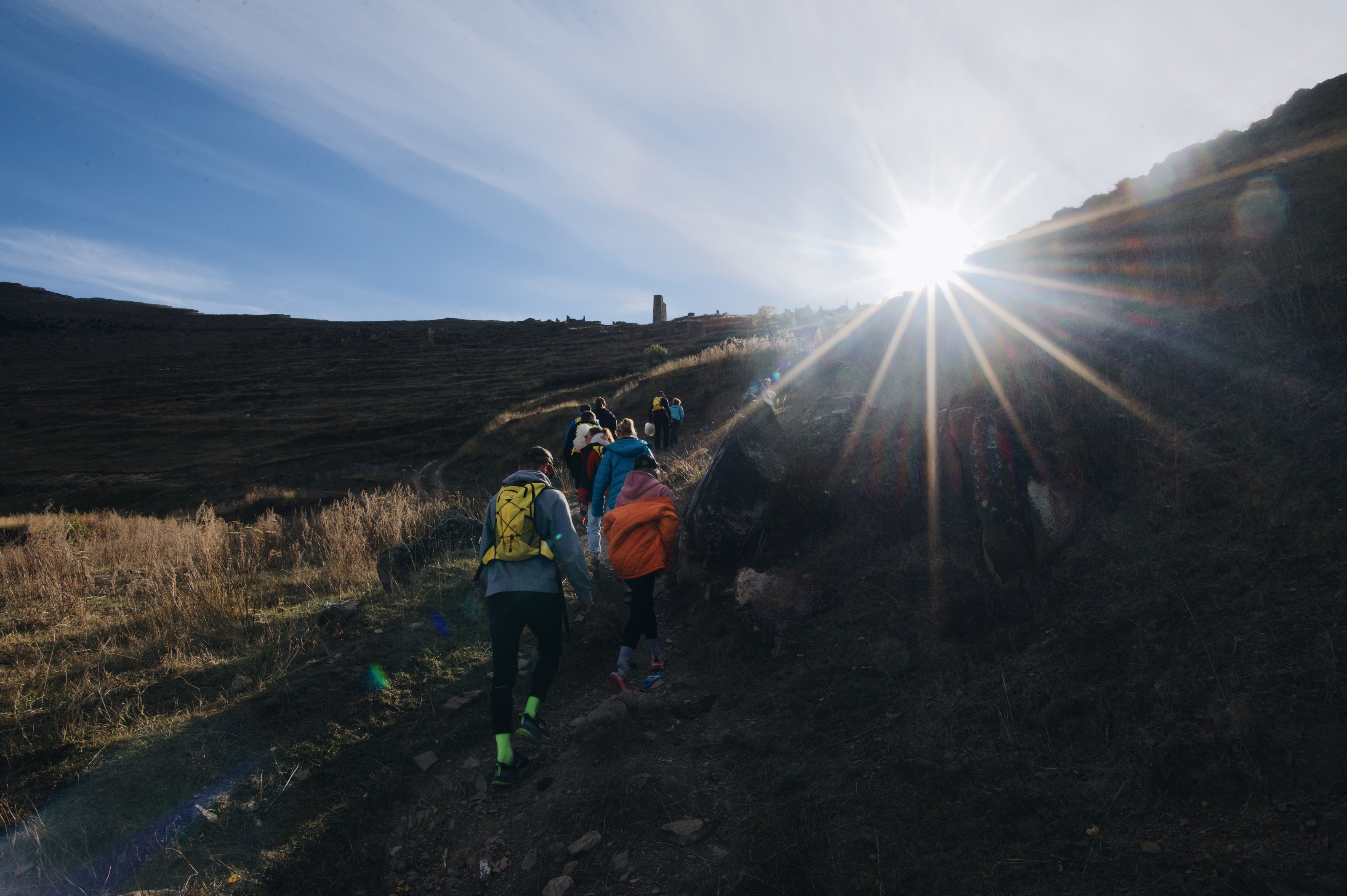 A group of trekkers with backpacks makes its way up a mountain trail in bright sunlight.