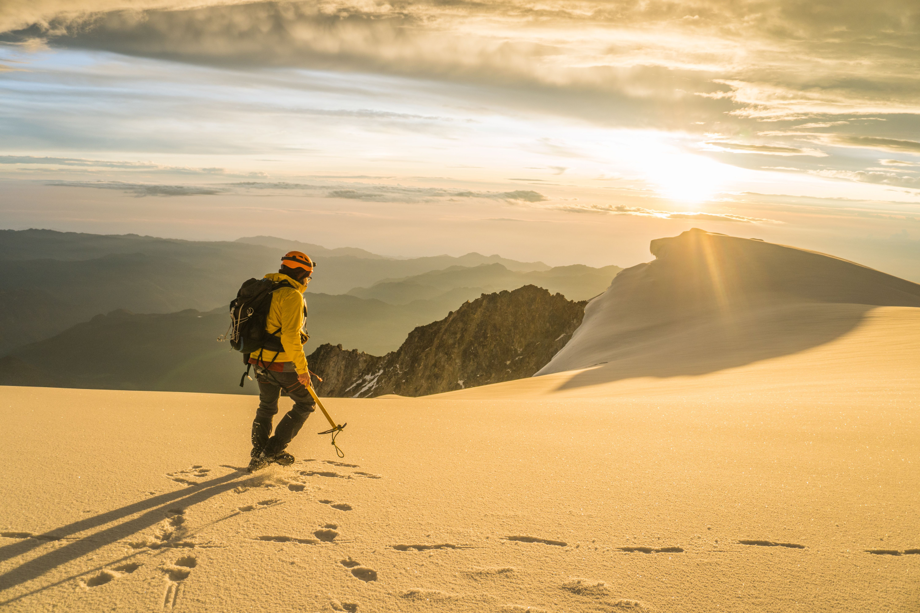 A lone mountaineer walking a snow-covered ridge under golden sunrise light.