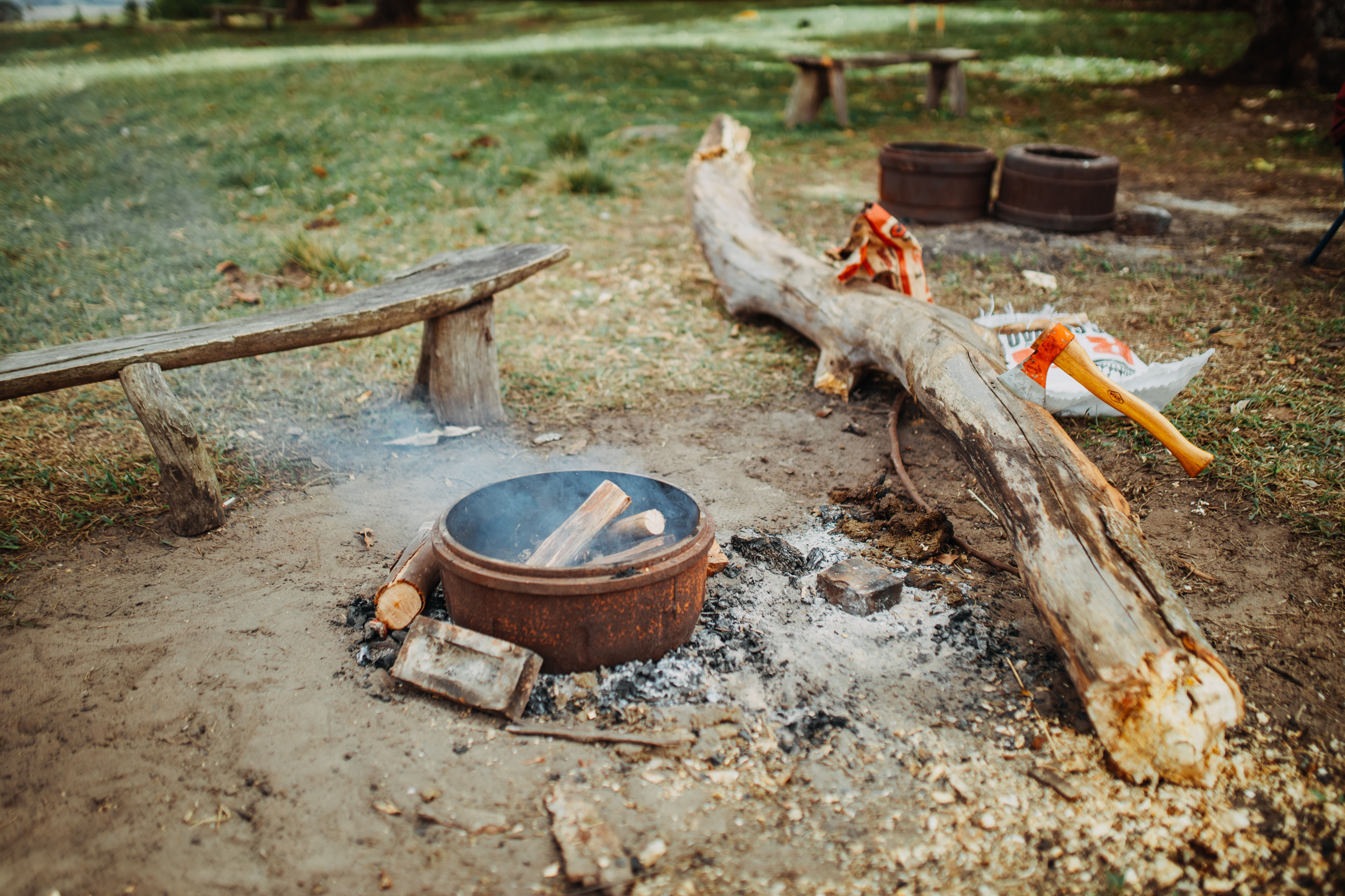 Simple tools and firewood are arranged beside a small campfire in a forest clearing.