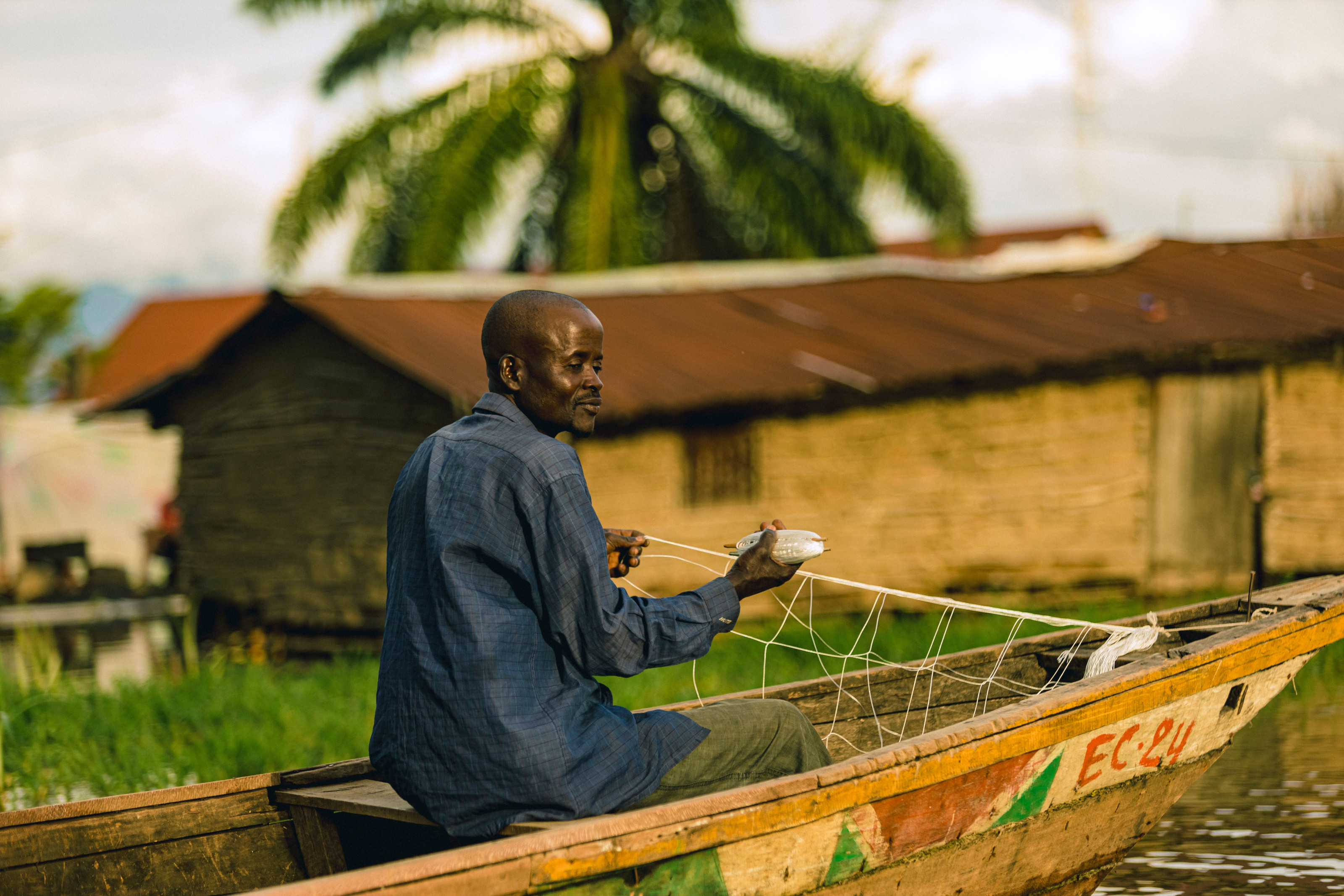 A man fishes from a small wooden boat near a riverside village of straw-roofed huts.