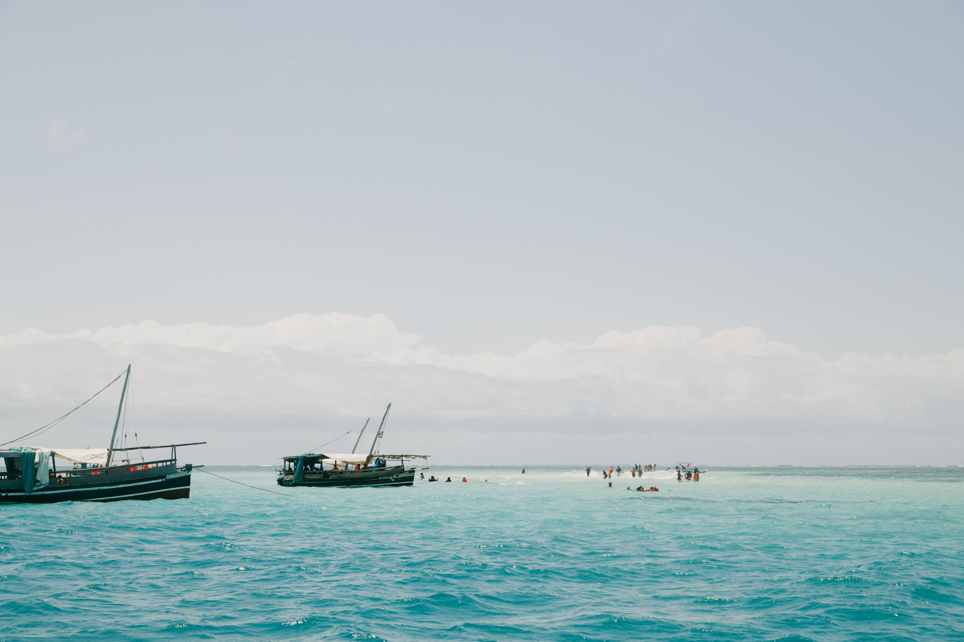 Traditional wooden boats float in crystal-clear turquoise coastal waters near a sandy shore.