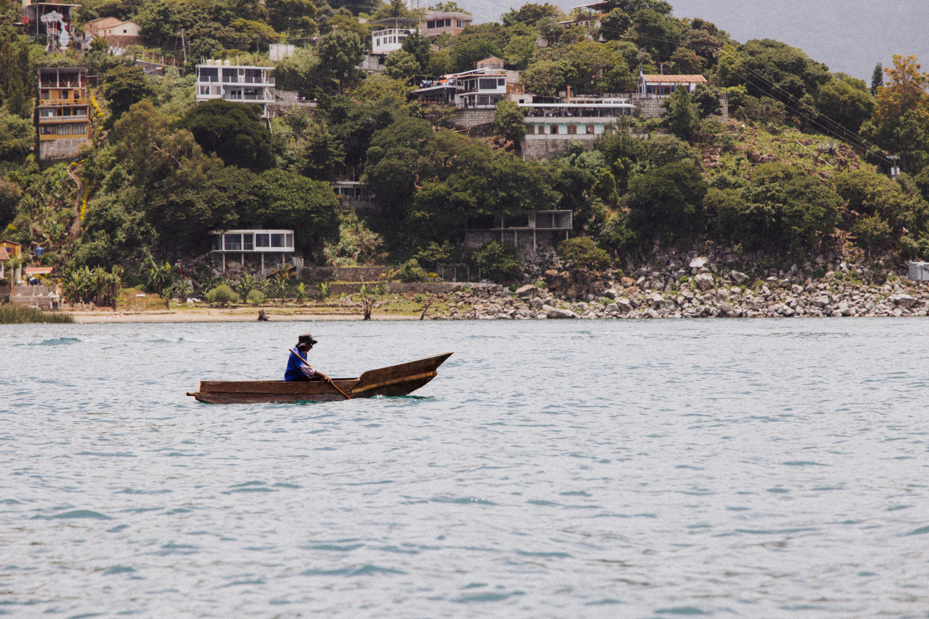 A wooden boat drifts near a lush hillside dotted with small houses above the water's edge.
