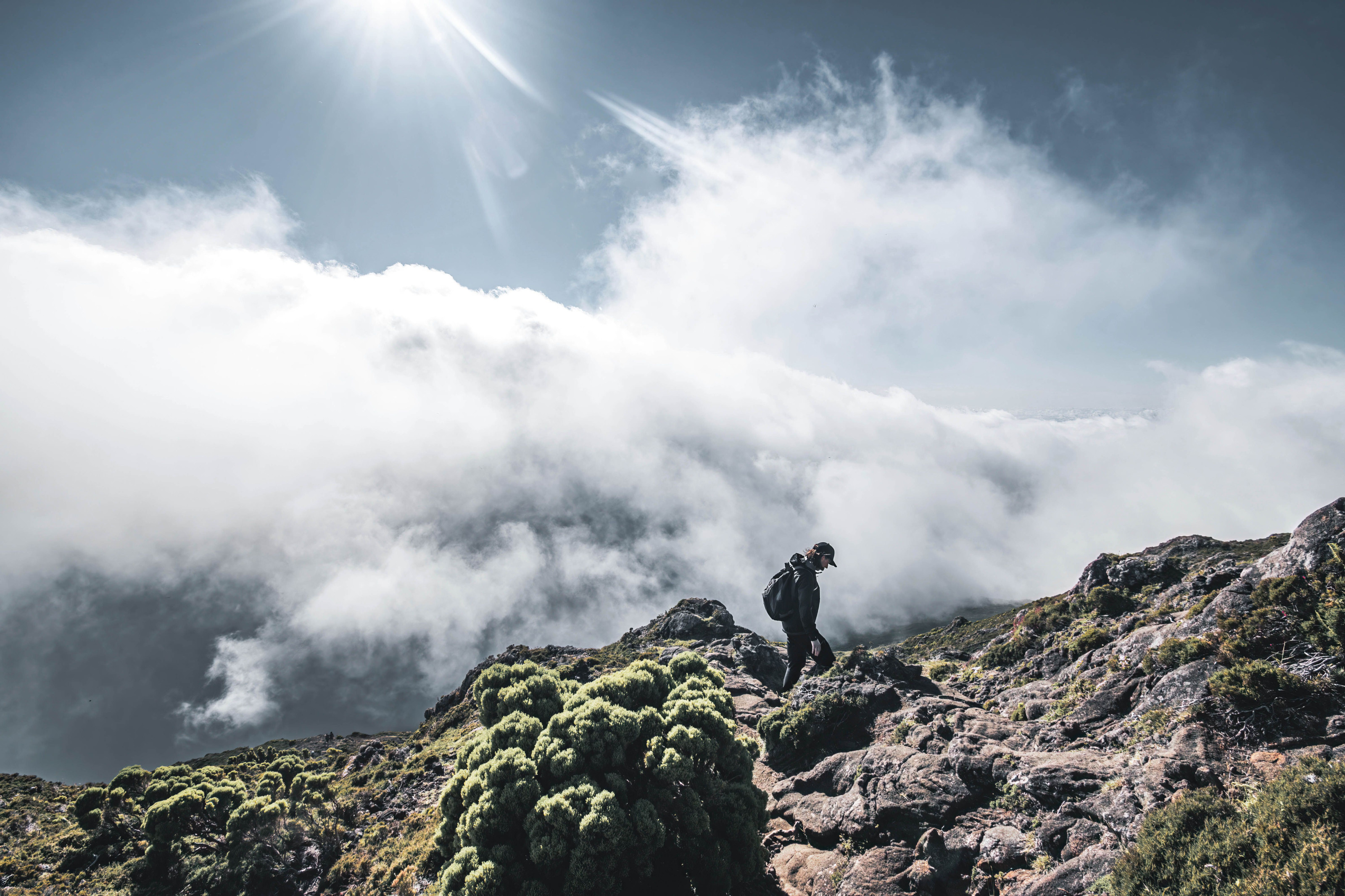 A lone hiker climbs a rocky mountain trail beneath a bright, cloud-streaked sky.