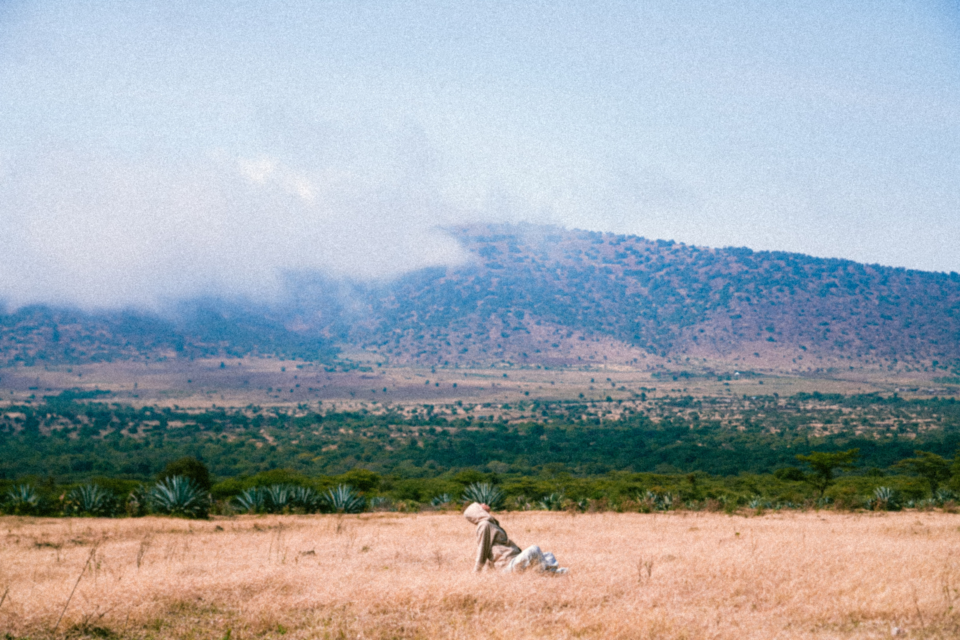 A lone figure walks across a vast, open African savanna beneath a wide, cloud-streaked sky.
