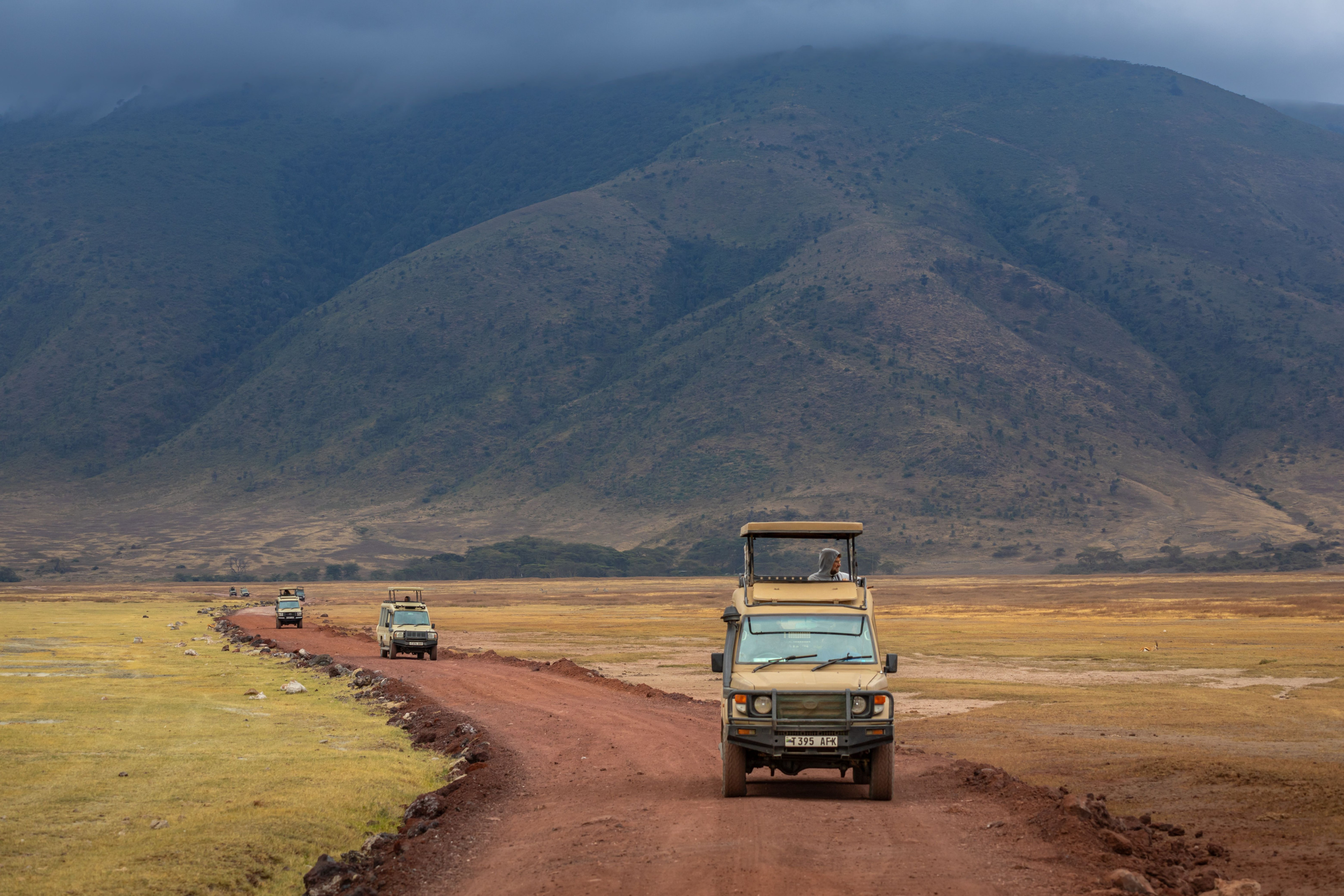 Safari jeeps kick up red dust on a crater-floor road beneath steep green walls.