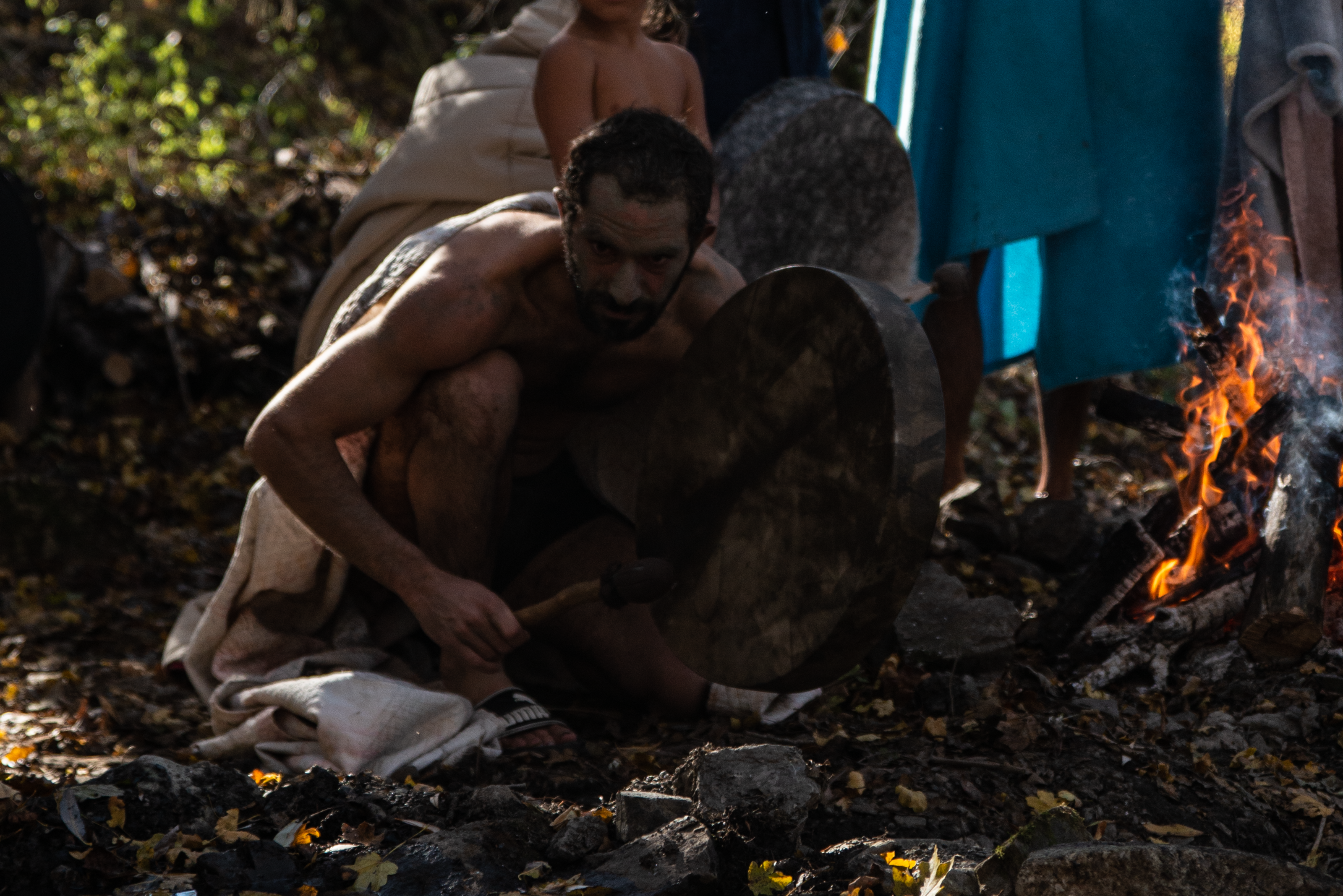A man plays a traditional drum beside a campfire as others gather nearby in the forest.