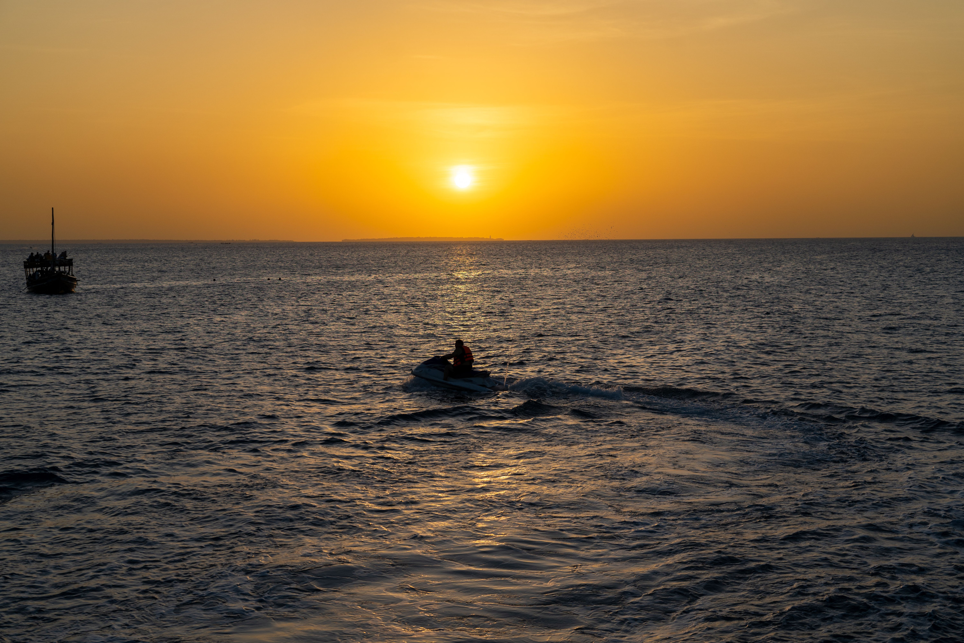 A small watercraft silhouette crosses a vivid orange ocean sunset.