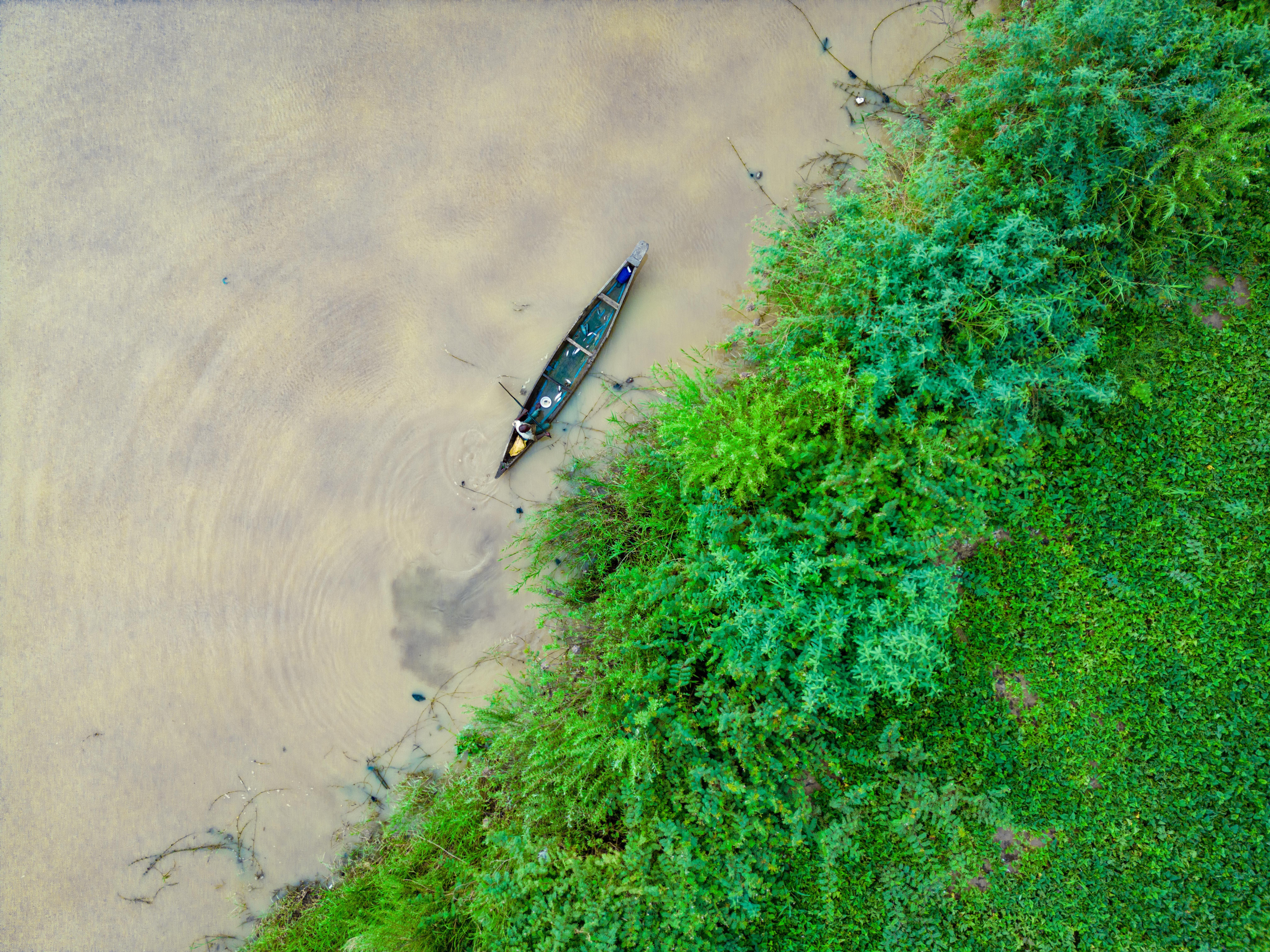 A fisherman paddles a dugout canoe along a lush, winding river, seen from above.