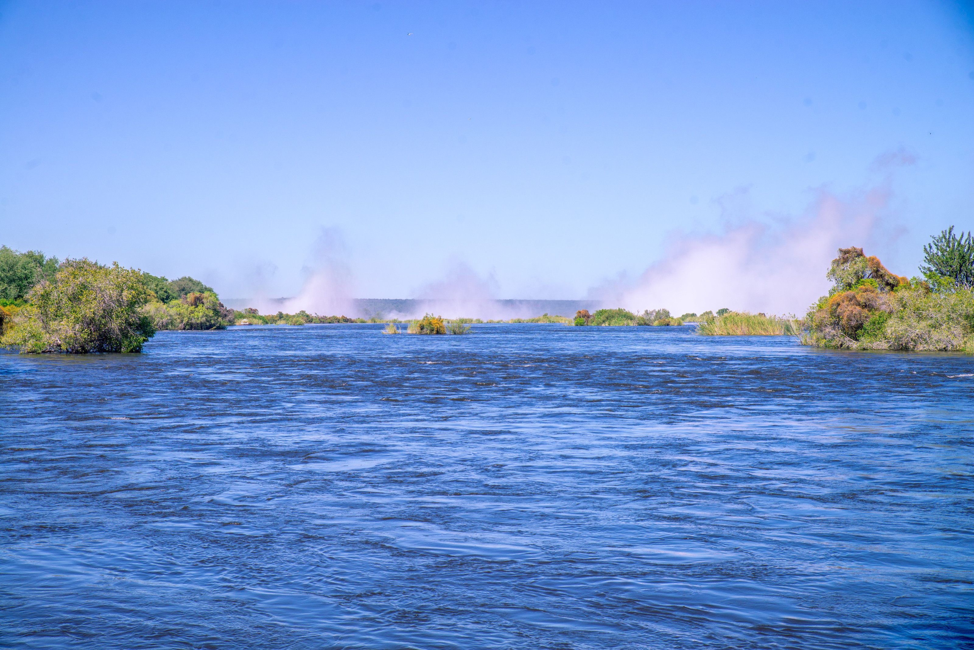 Mist rises off a wide river winding through a green gorge under a clear sky.