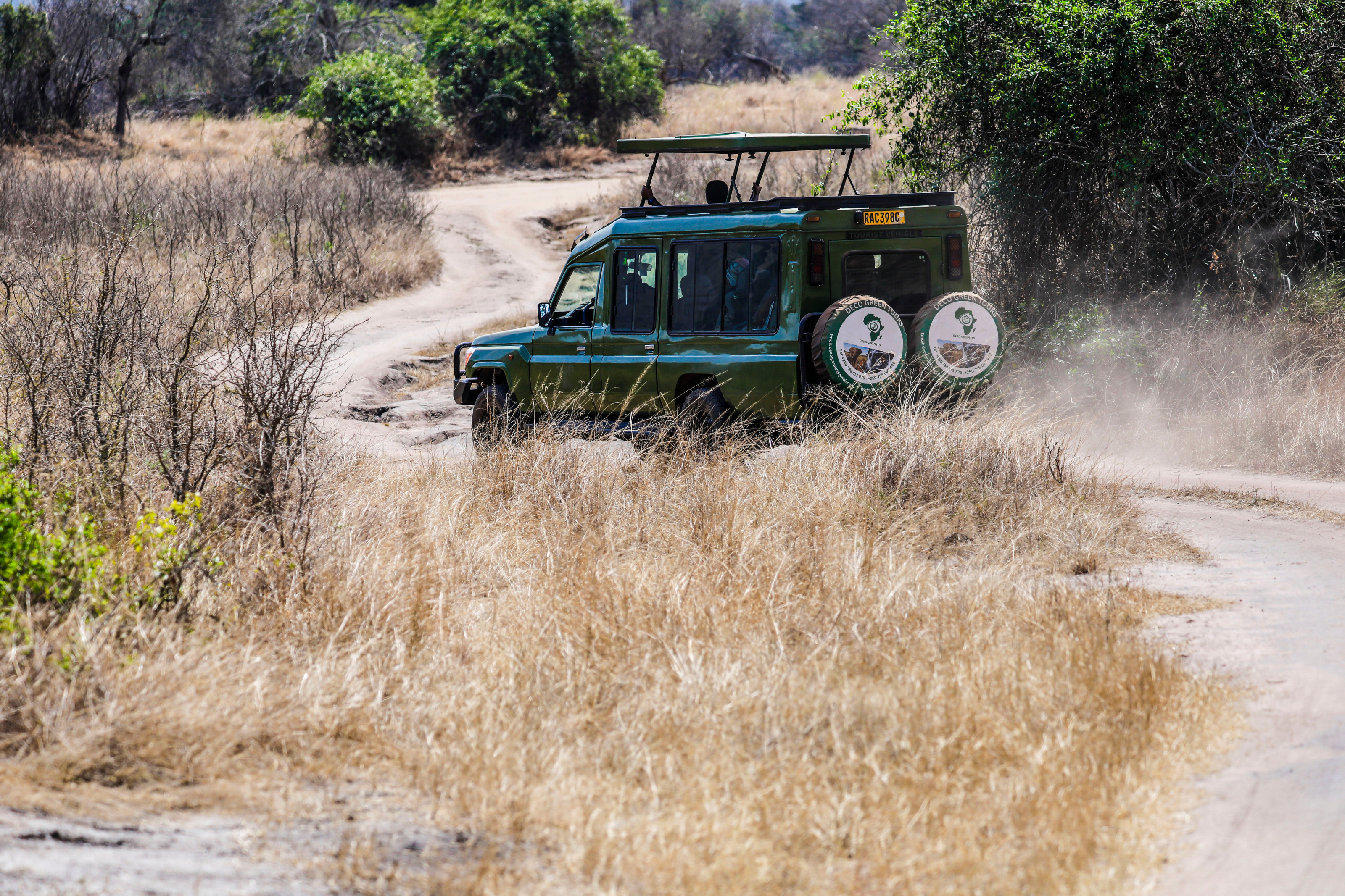An open-sided safari vehicle kicks up dust as it travels along a dirt track through golden East African savanna.