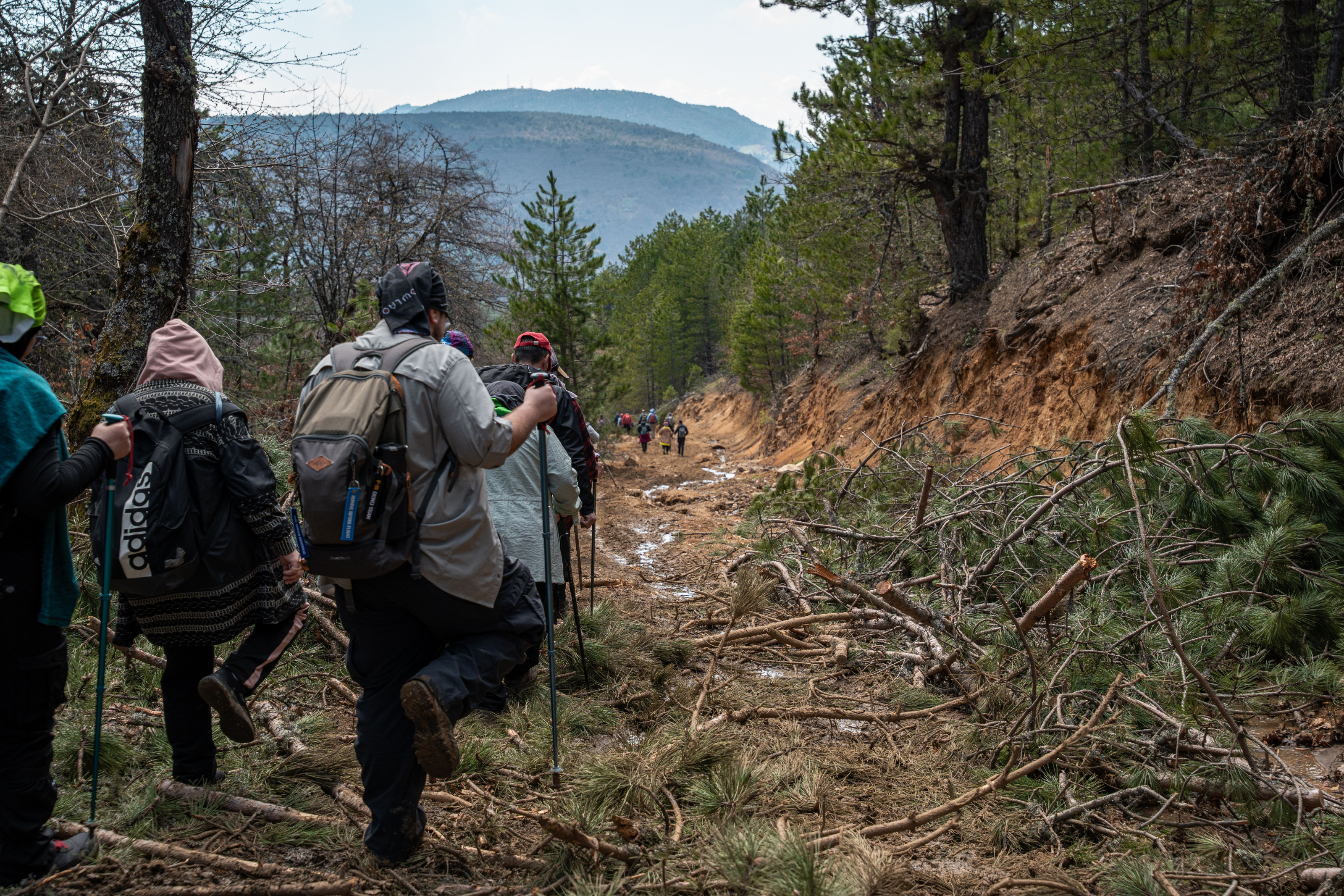 A line of hikers pushes along a muddy forest trail through dense, mountainous greenery.