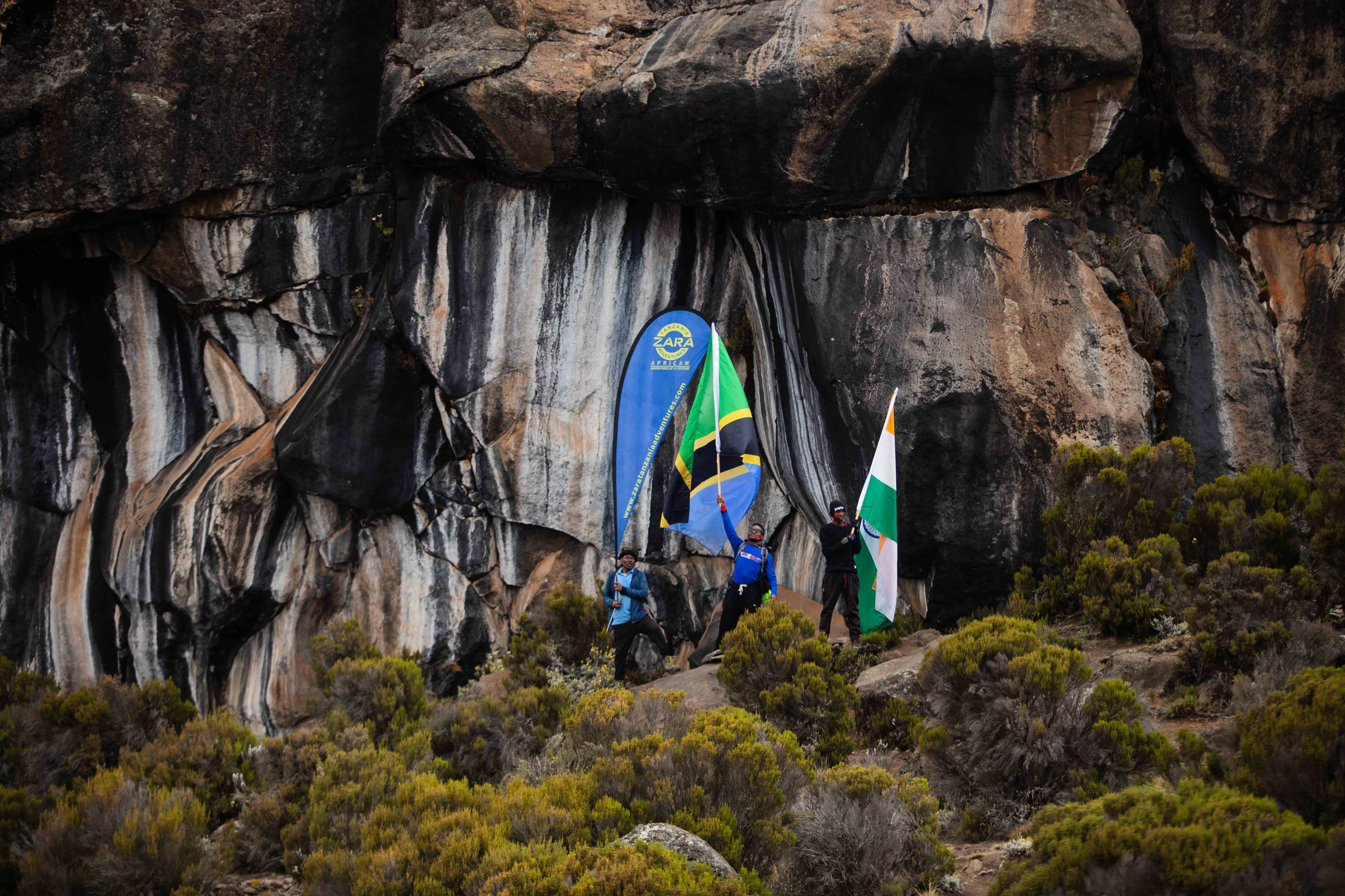 Hikers pose with flags at a rocky cave entrance partway up Kilimanjaro.