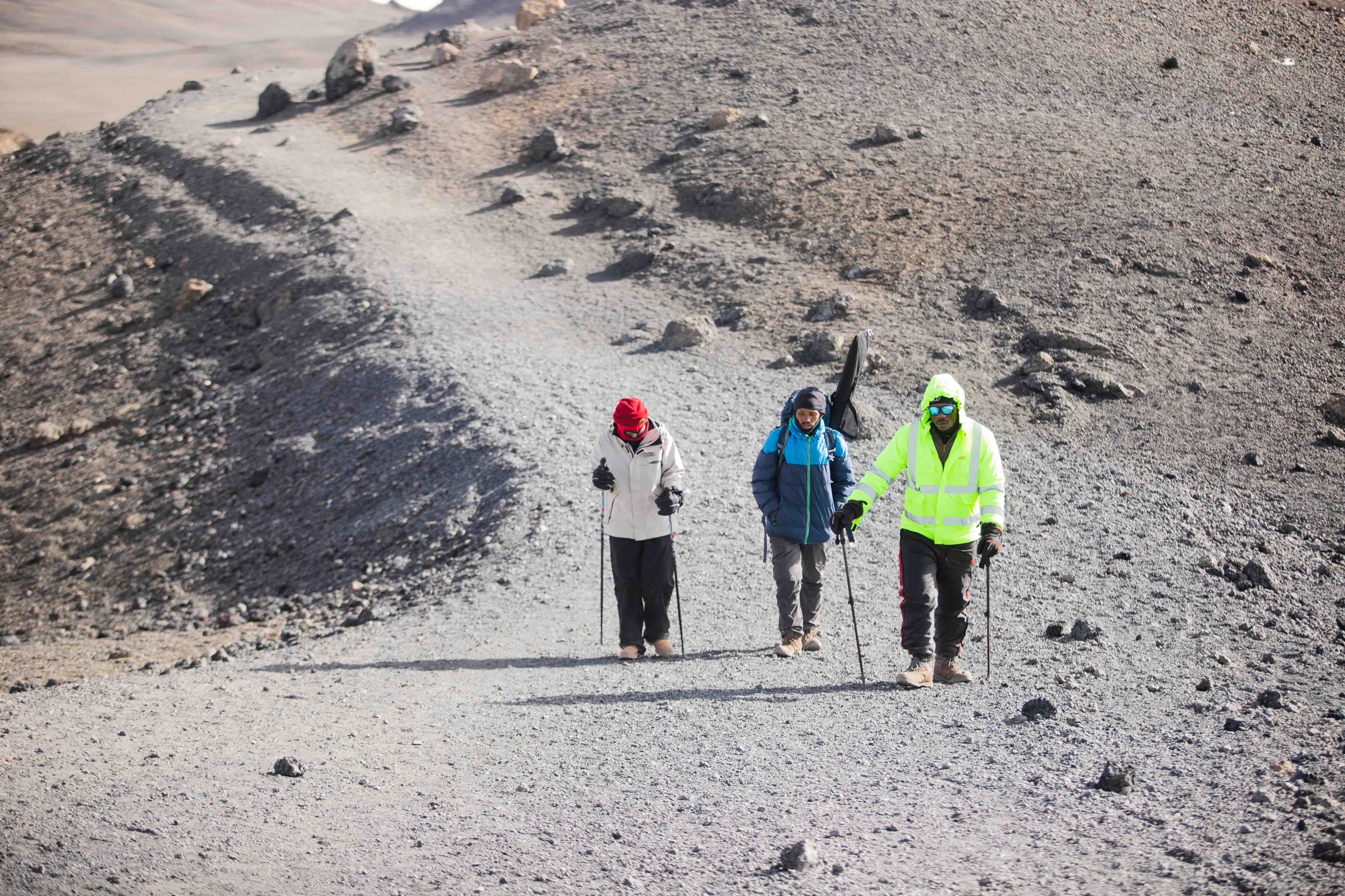 Three hikers with trekking gear making their way along a rocky volcanic slope.