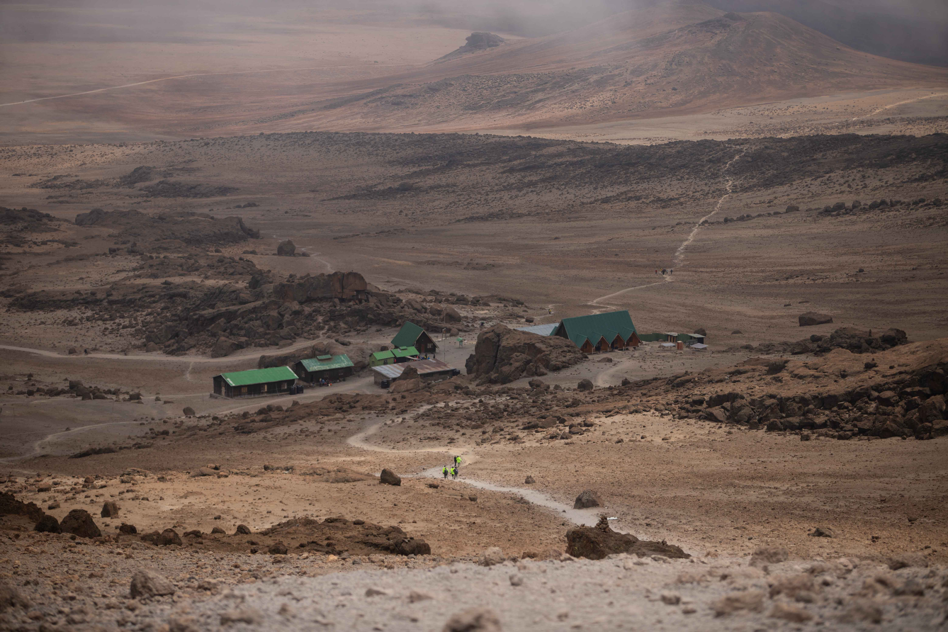Trekkers approach a remote mountain camp across a wide, rugged high-altitude plateau.