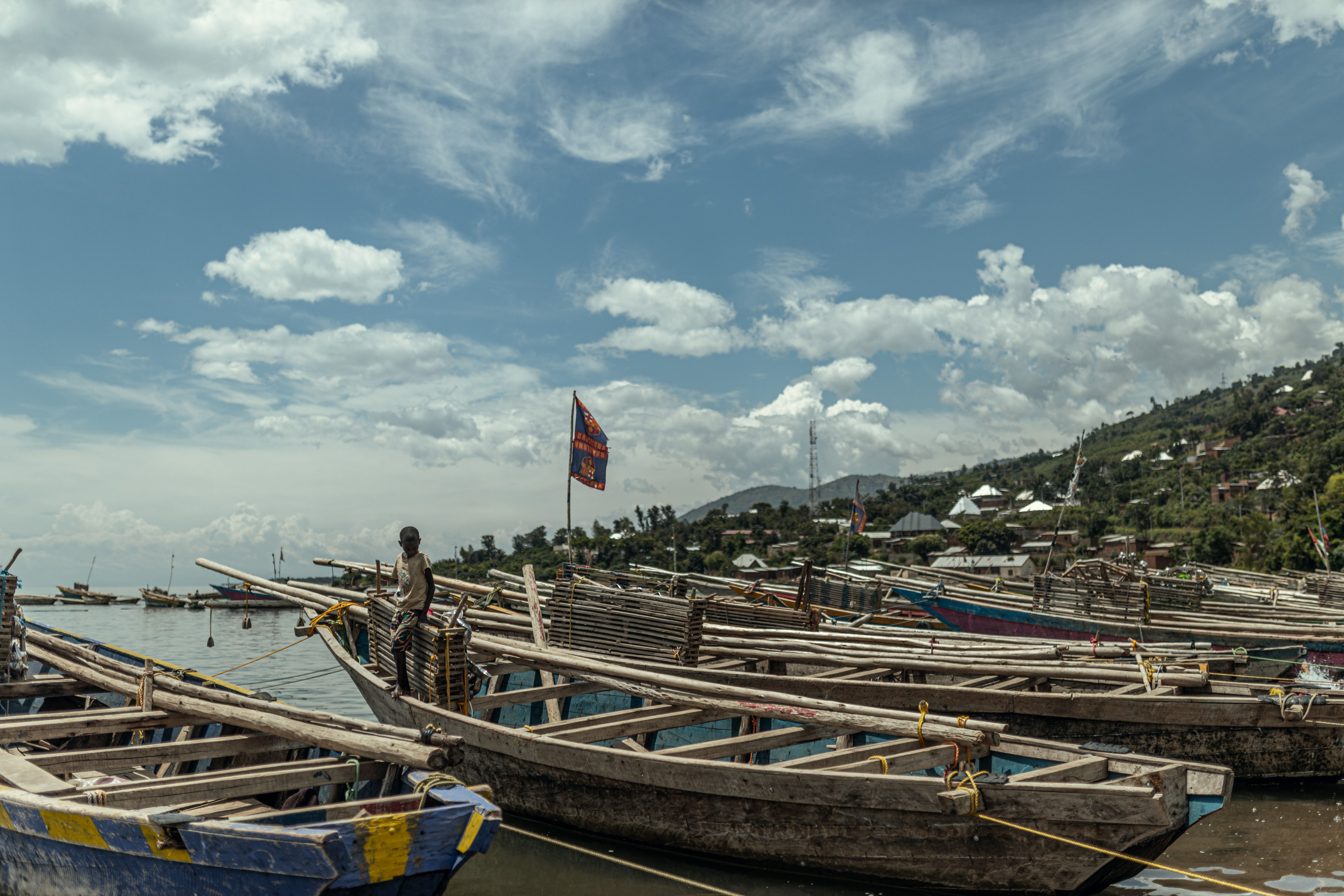 Wooden fishing boats sit moored along a quiet lake shore beneath distant rolling hills.