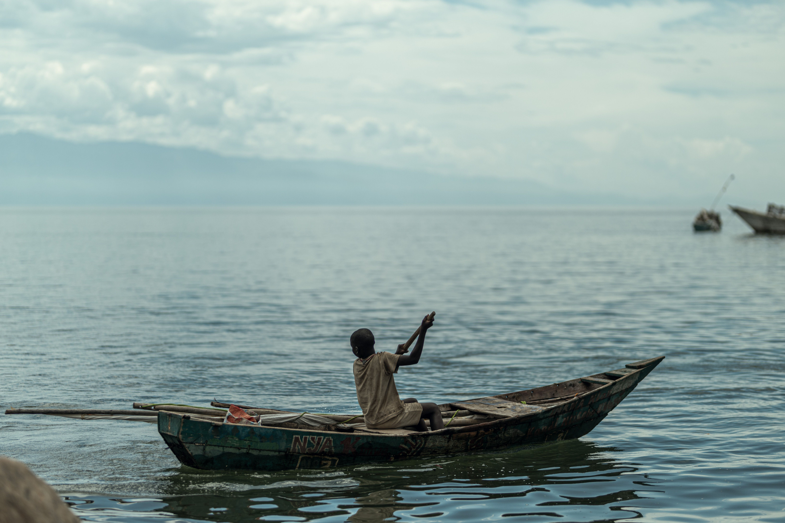 A lone fisherman paddles his wooden boat across still water under an overcast sky.