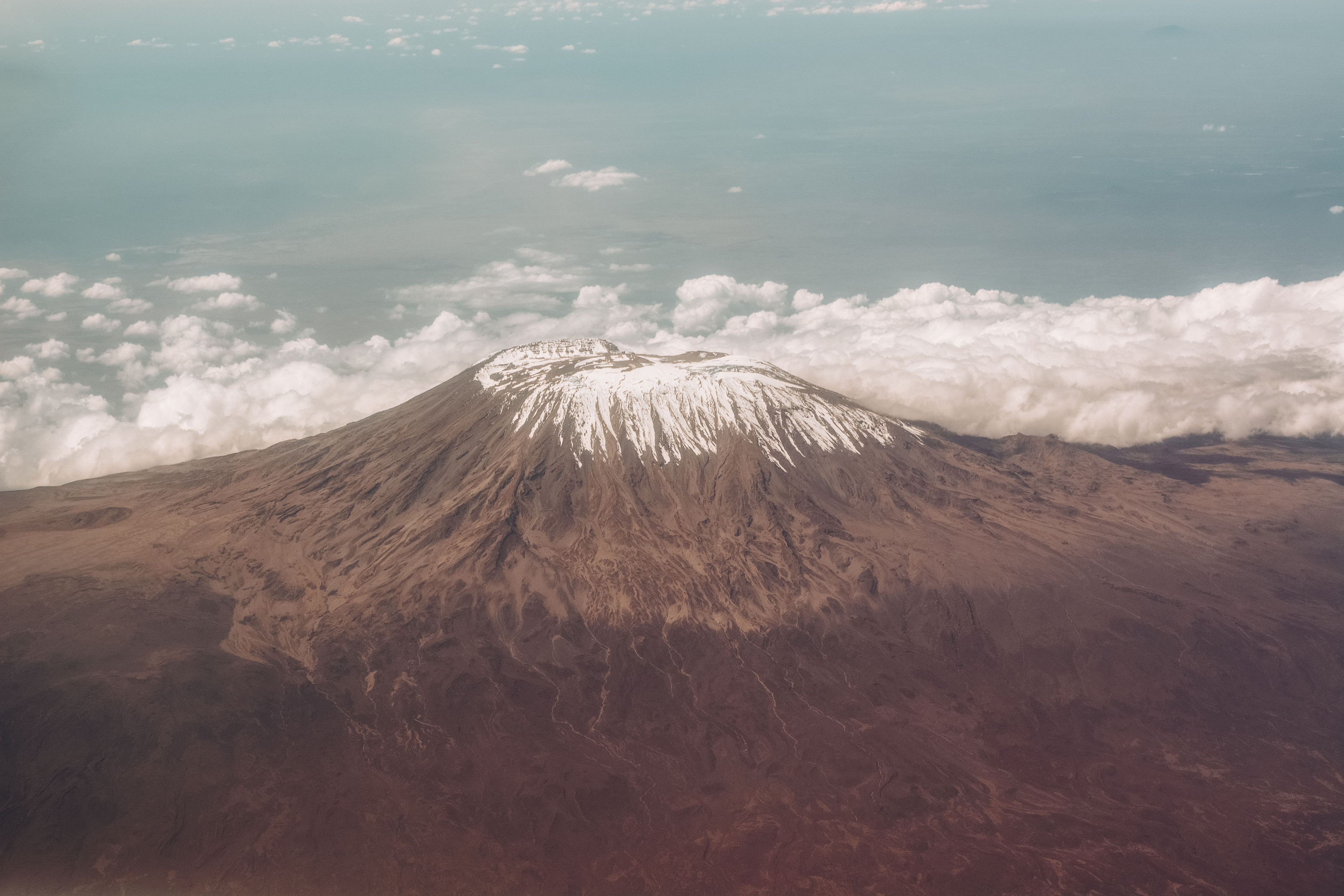 Clouds drift below the snow-capped summit of a towering volcanic peak seen from above.
